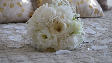 Disparo-Giratorio-Y-Enfoque-En-El-Ramo-De-Flores-Blancas-De-La-Novia-En-Una-Cama-Lujosa-Con-Pétalos-De-Flores-Blancas-Por-Todas-Partes,-Ropa-De-Cama-Blanca-Y-Dorada