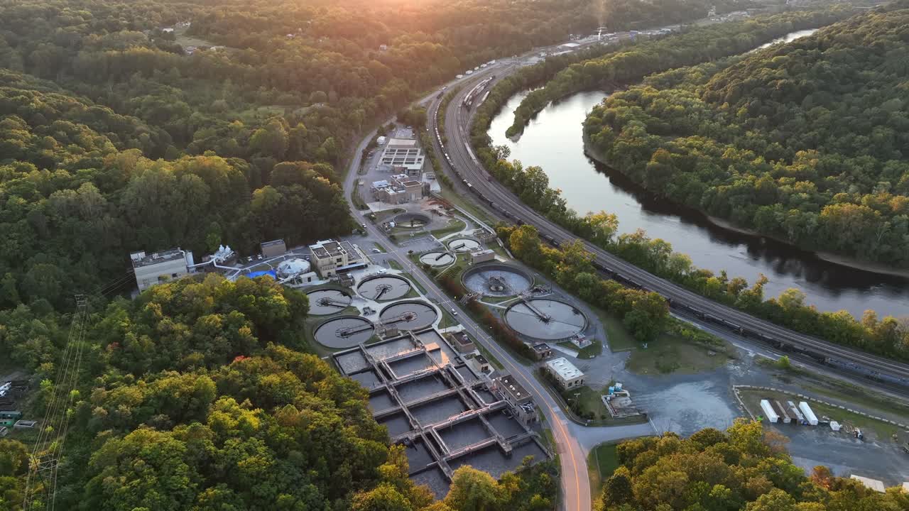 Traffic on road between wastewater treatment factory in USA. Sunset time in summer. Tranquil river in forest landscape of town. Aerial top down shot