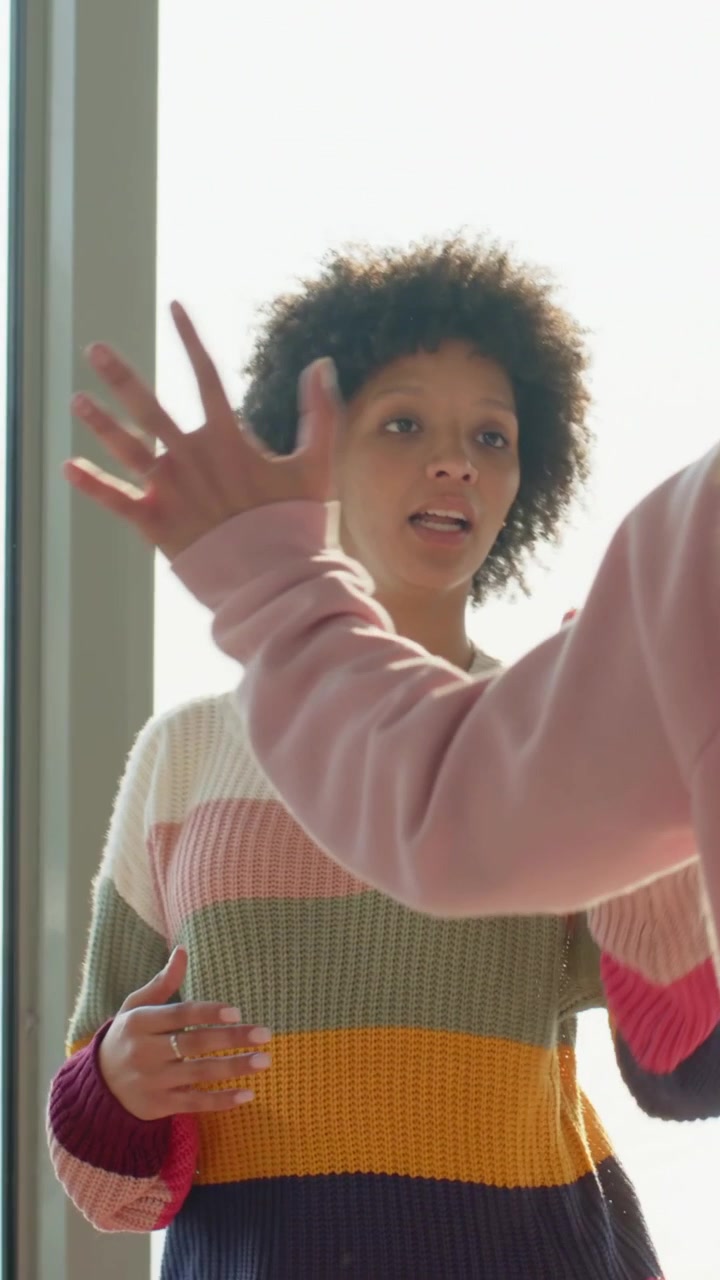 Vertical video: Smiling pink-hoodie woman stepping by glass, reaching striped friend, greeting