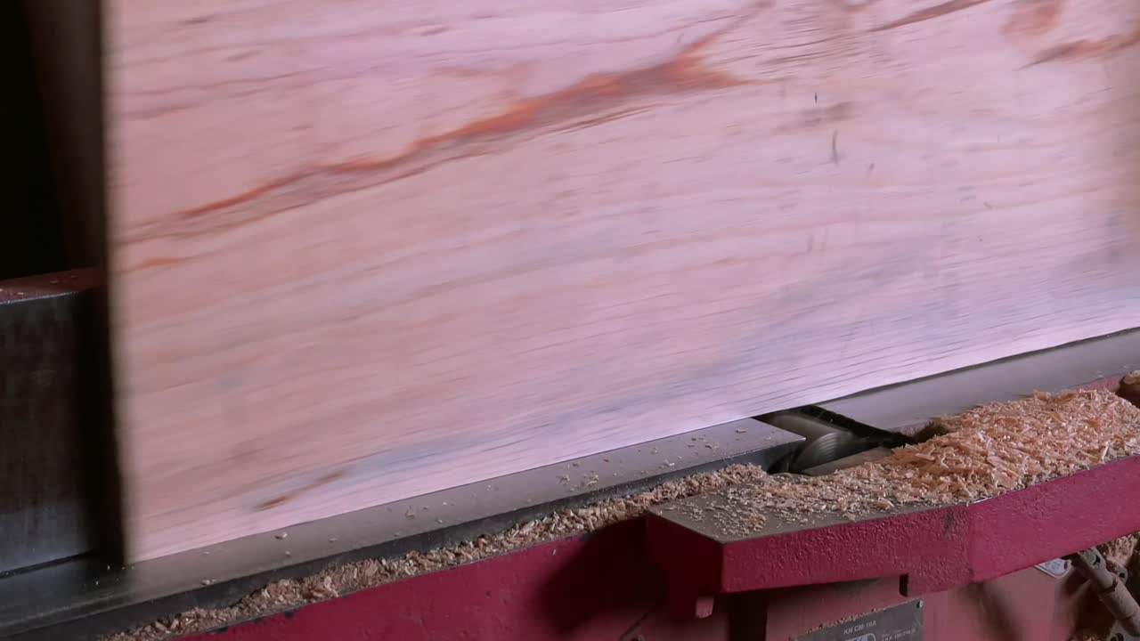 Wood board with natural grain is being planed on a jointer machine with visible shavings near a carpentry workshop. Static close-up shot