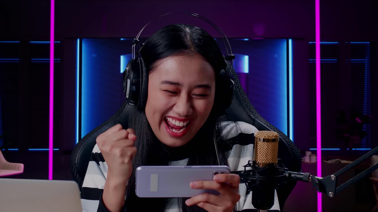 primer plano de una feliz mujer asiática jugadora con auriculares y computadora celebrando la victoria en el juego de teléfono móvil en una habitación iluminada por luces de neón en retro