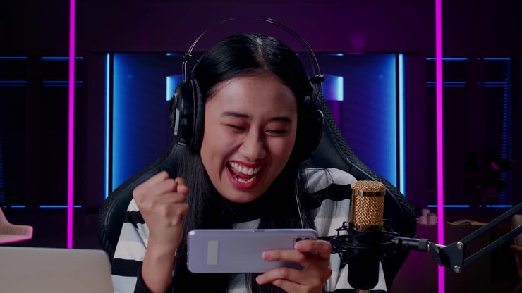 Close Up Of Happy Asian Woman Gamer With Headphone And Computer Celebrating Winning The Mobile Phone Game In Room Lit By Neon Lights In Retro