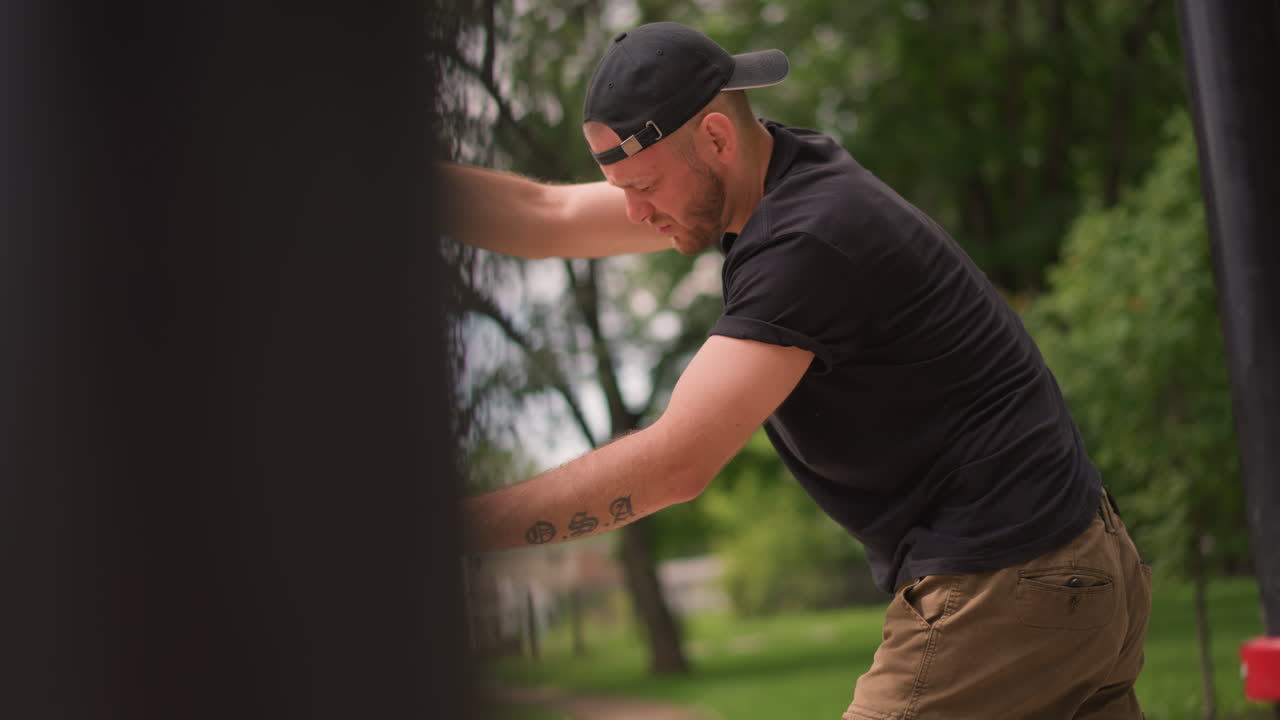 White Man Aligning Metal Pole Outdoors With Focused Effort, Hands Gripping Black Post, Cap Worn Backwards, Concentrated Stance In Leafy Park Setting, Daytime Training Or Maintenance Vibe