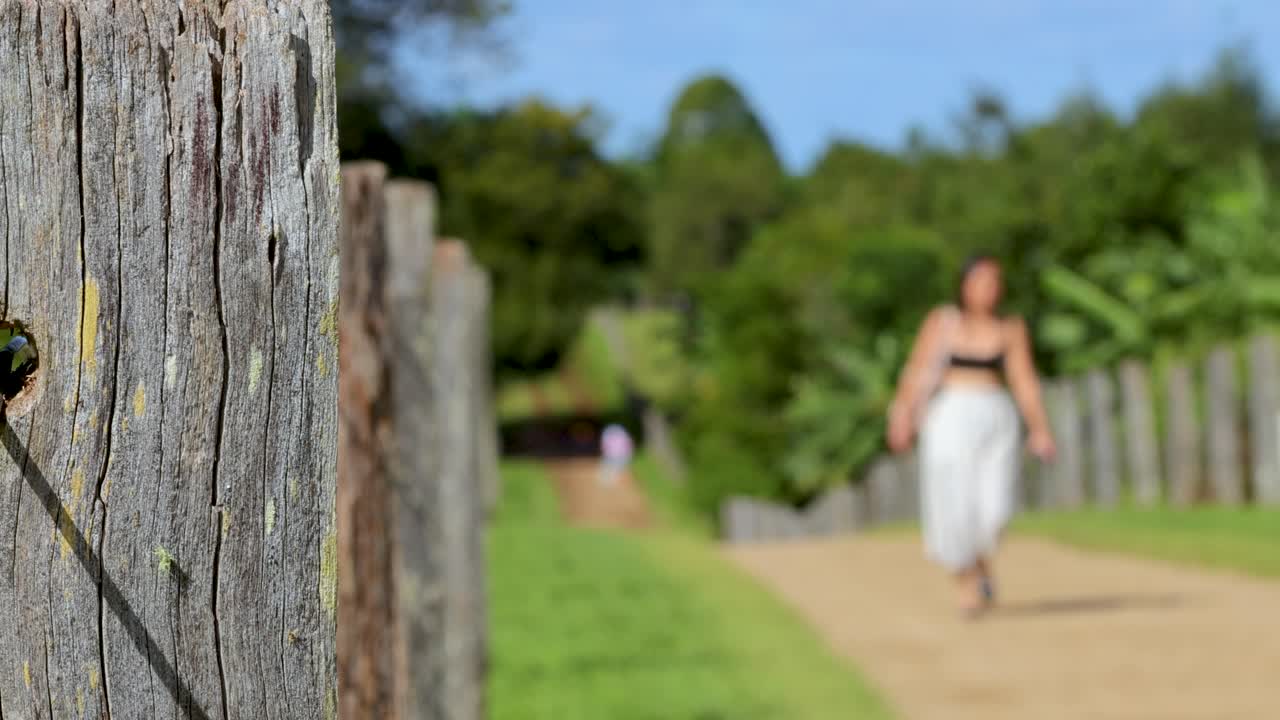 A woman in a white skirt walks down a sunlit dirt path next to a rustic wooden fence, surrounded by lush greenery. Shallow depth of field, steady camera