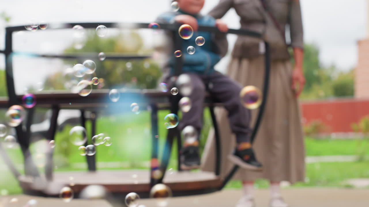 Close up of vibrant bubbles drifting through air while woman stands behind blurred merry-go-round structure gently turning child, playful atmosphere under cloudy sky in green outdoor park setting