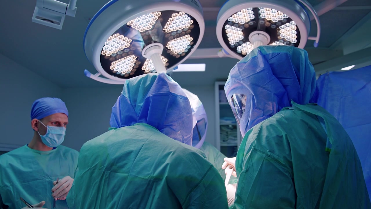 Vista posteriore di due medici che indossano camici e caschi durante un intervento chirurgico. Lampade rotonde e luminose sono sopra i medici.