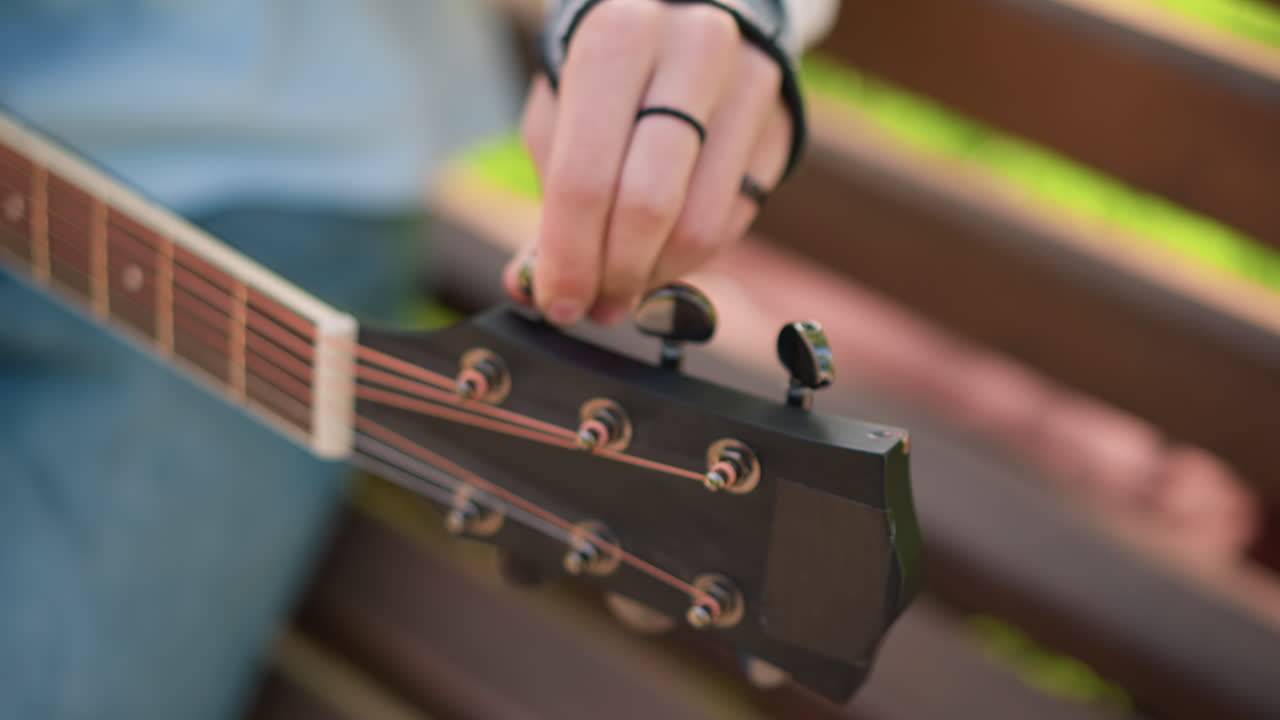 closeup acoustic guitar tuning on bench, young guitarist adjusting tuning pegs with ringed fingers, denim jeans visible, sunlight dappling wood, relaxed introspective practice session focused