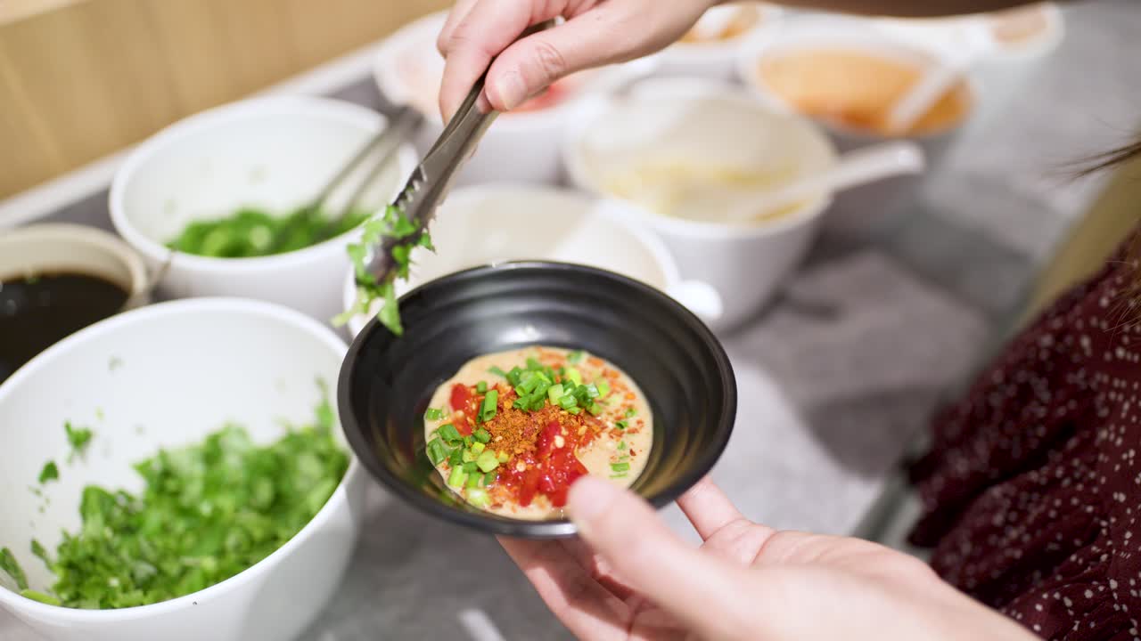 Hand adds cilantro, chili, and green onion to dipping sauce in bright buffet setting