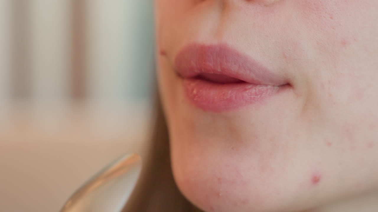 Elegant young lady dips tablespoon of hot chocolate drink into mouth, slowly bringing it out with satisfied expression, focus on lips and spoon with soft indoor background in warm lighting ambiance