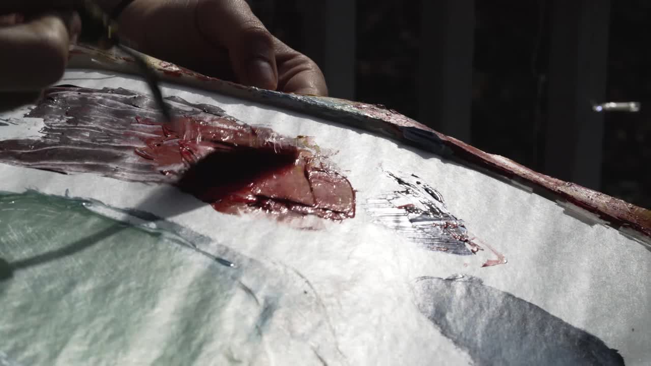 Female artist mixes yellow with red paint on her palette outside at sunset with palette knife, camera pans right