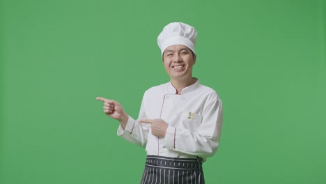 el chef asiático sonriendo y señalando al lado mientras está de pie en el estudio de fondo de la pantalla verde