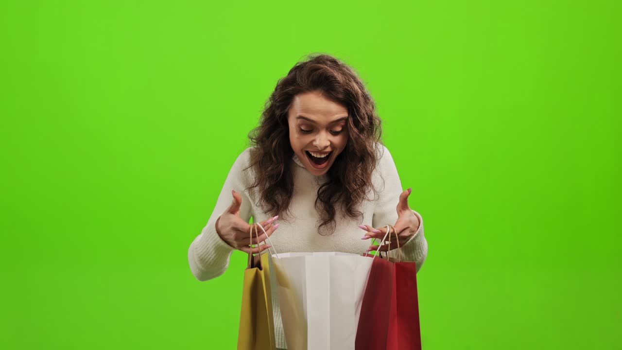 한 여자가 쇼핑 가방을 들여다보고, 미소 짓고 기하고, 머리를 들고 웃고, 초록색 바탕에 서 있다.