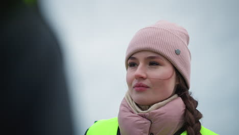 young female worker in warm winter clothing, pink knit hat, and bright reflective vest standing outdoors on cold cloudy day, speaking with attentive expression, possibly engaged in discussion about work