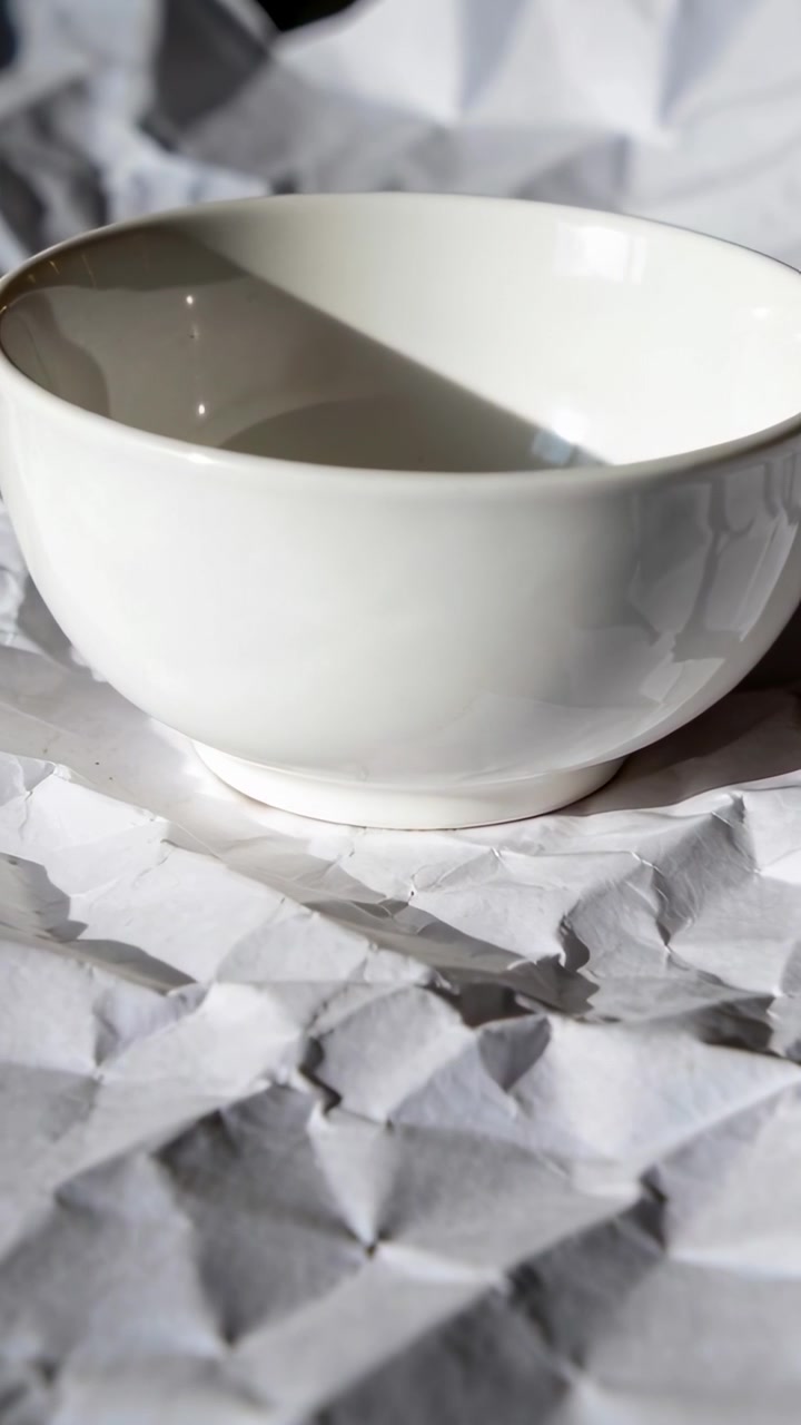 Camera moving closer, framing white ceramic bowl on crumpled paper, revealing shadow for product ad