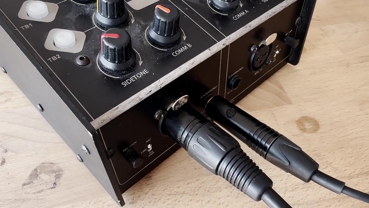 Primo piano di un pannello di controllo di un mixer audio con manopole colorate, pulsanti cue e indicatori di livello di uscita su un tavolo di legno.
