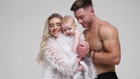 happy family of three in the studio. lady waves her child and father kisses his baby boy. white backdrop.