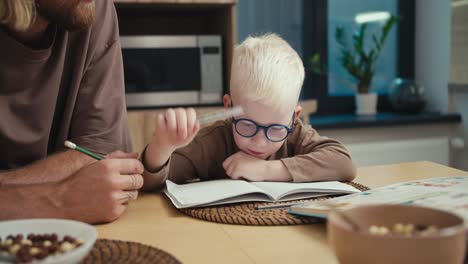 Close-up-shot-of-a-little-albino-boy-with-white-hair-wearing-blue-glasses-puts-down-his-pen-and-raises-his-two-palms-up-to-count-on-his-fingers-during-his-math-homework