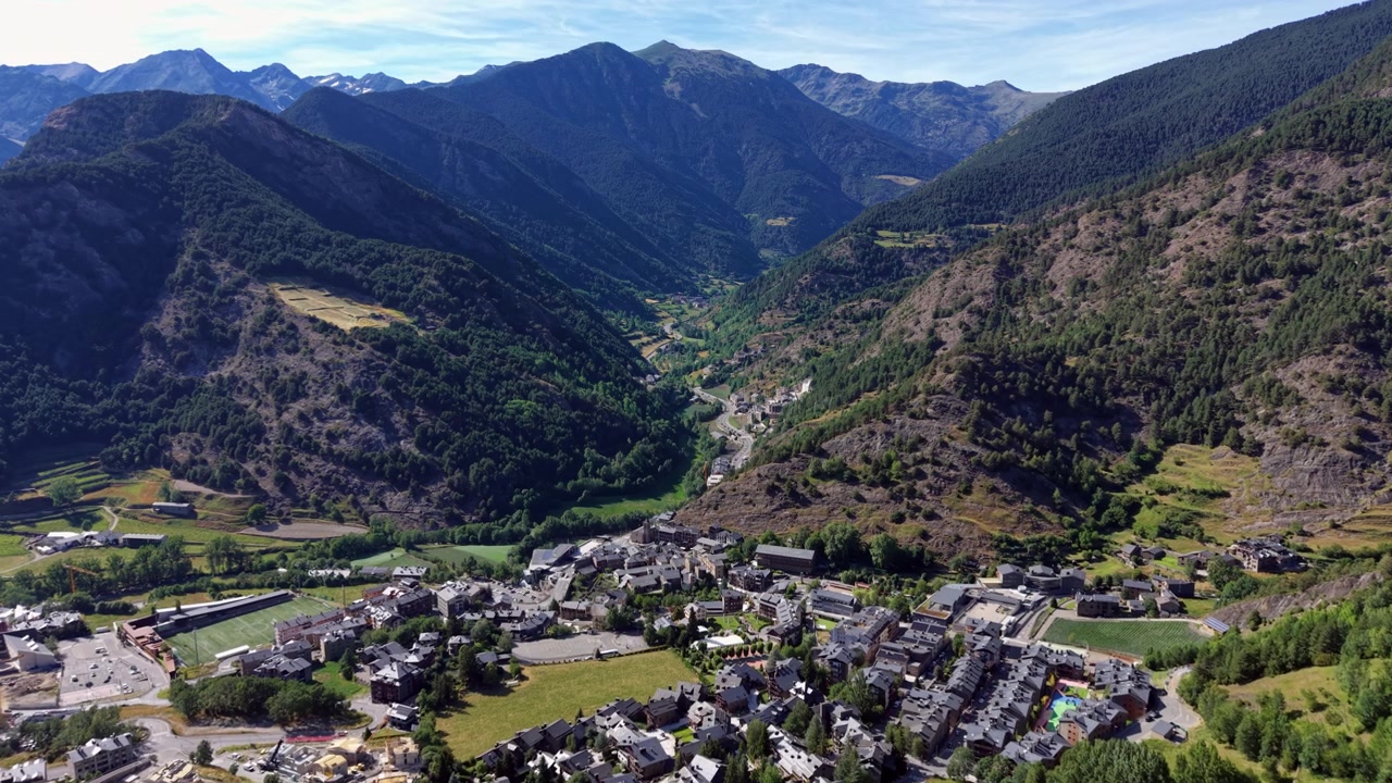 Il villaggio di Ordino immerso in una lussureggiante valle pirenaica ad Andorra, circondato da pendii montuosi boscosi, campi verdi e strade tortuose sotto un cielo estivo sereno.