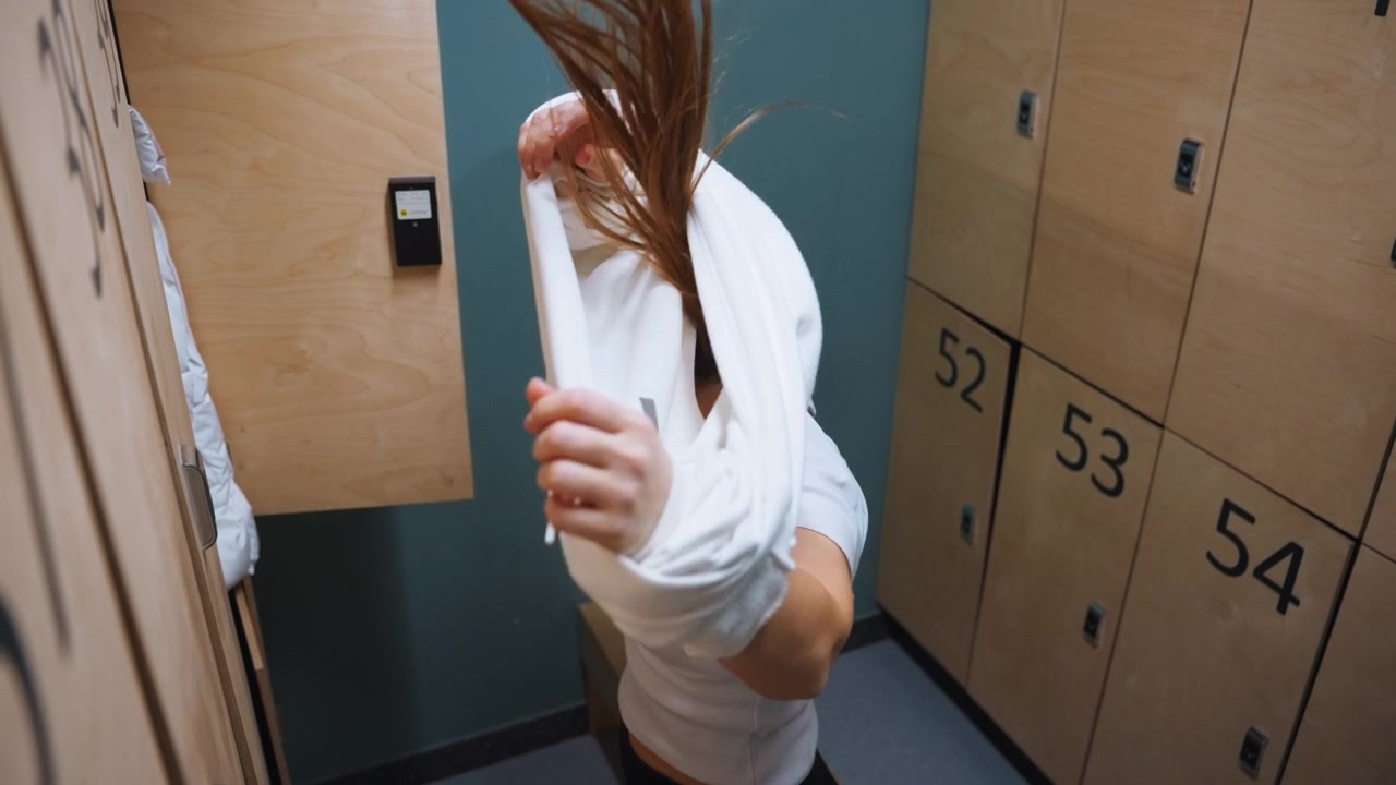 White woman in locker room changing, removing warm sweatshirt after workout, focused calm expression, numbered wooden lockers and teal wall, candid overhead angle, preparing post workout routine