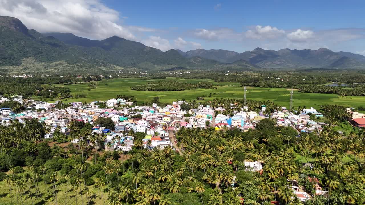 Sweeping drone footage of an expansive town Courtallam Tamil Nadu, set across a hilly landscape. The settlement is characterized by houses interspersed with large patches of green farmlands