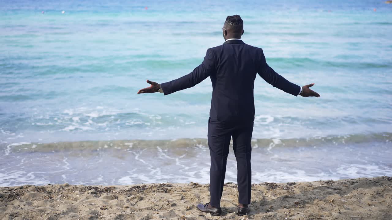 Back view of happy excited African American businessman stretching hands admiring beauty of turquoise azure foamy waves rolling on sandy beach in slow motion. Relaxed tourist enjoying vacations.