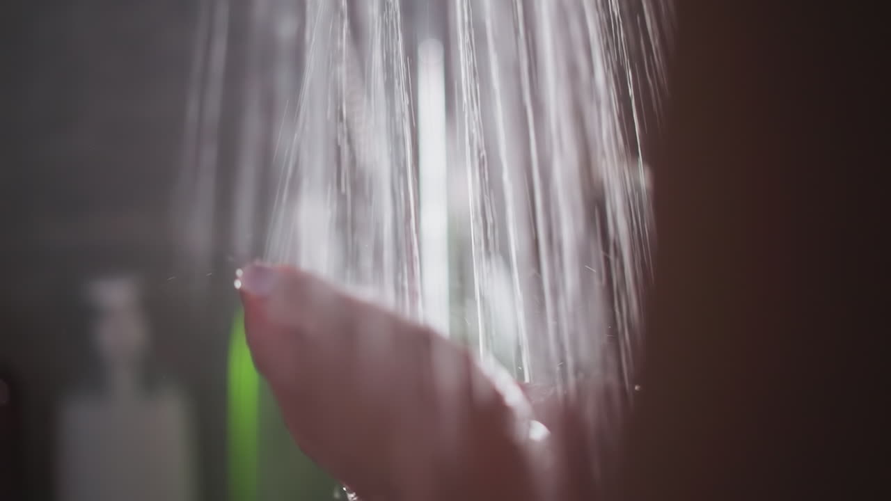 여성은 뜨거운 물의 끊임없는 폭우 아래 서 있습니다. 여자는 샤워에서 은 물의 스프레이 아래 손을 움직입니다. 여성의 손은 흐르는 하천에서 바람을 피우고 있습니다.