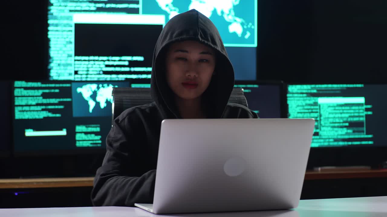 Asian Female Hacker In A Hood Looking At Camera And Crossing His Arms While Works On A Laptop With Maps And Data On Display Screens Of Multiple Monitors In A Dark Office Room