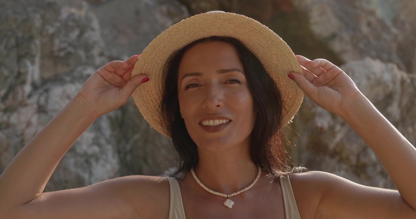 Donna sorridente con cappello di paglia e costume da bagno su una spiaggia rocciosa