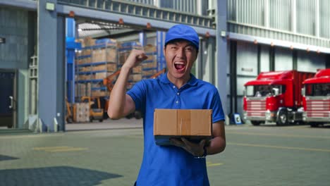 asian male courier in blue uniform celebrating succeed delivering a carton, outside of logistics distributions warehouse