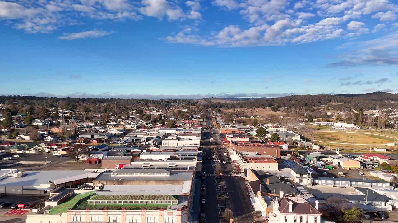 Drone camera glides above a sunlit main street in Coonabarabran, New South Wales, revealing urban buildings, traffic, and residential areas under blue skies