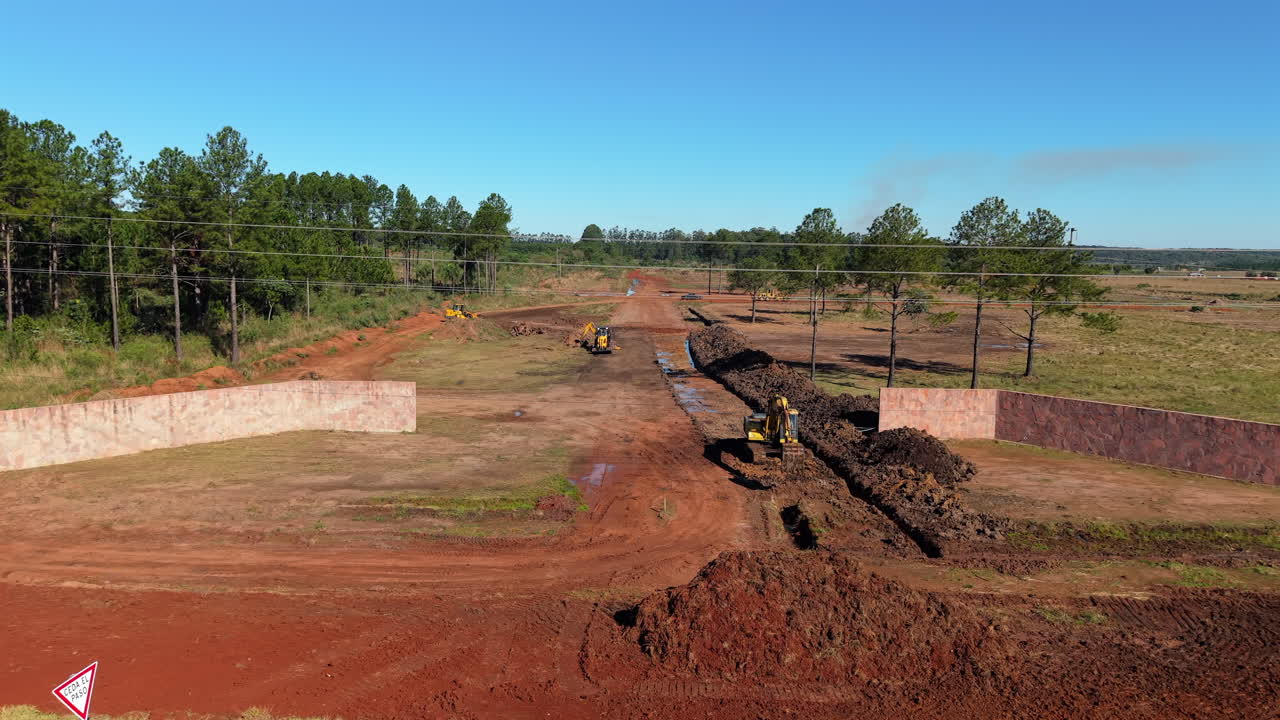 Excavators digging wide trench in red soil field under clear blue sky, Argentina