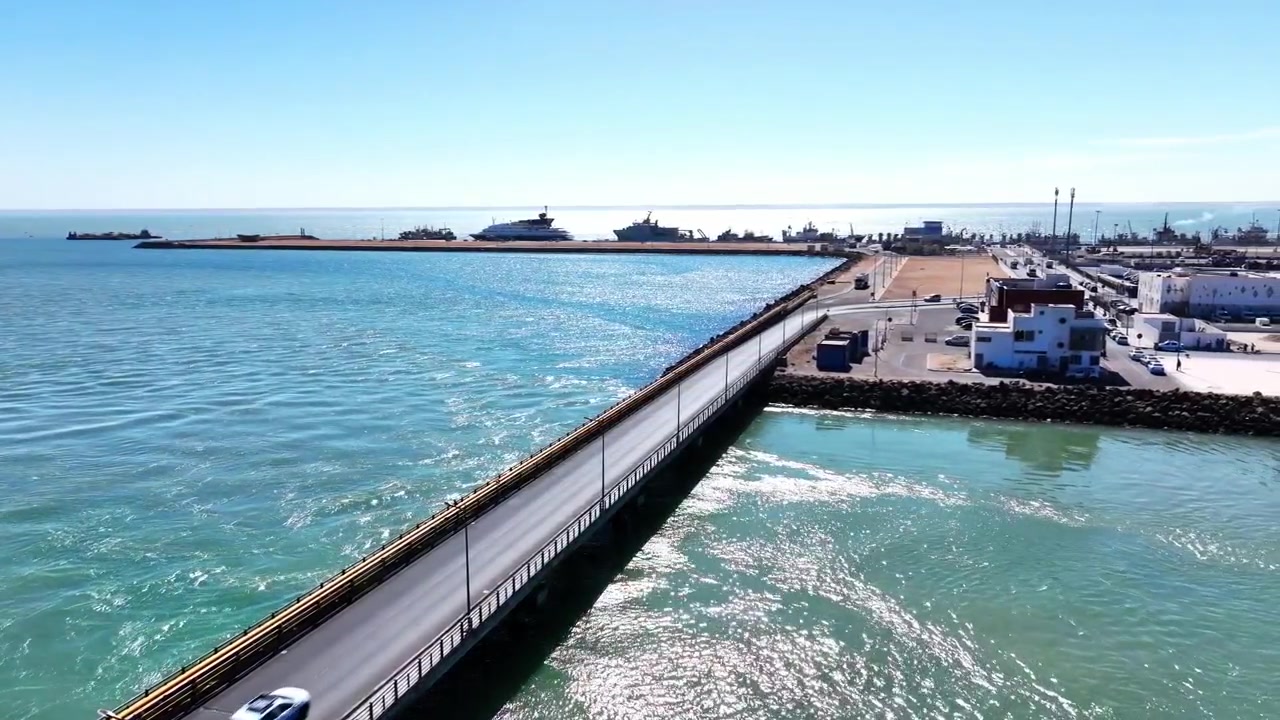 Vista aérea de dron de una calzada marina cruzando aguas turquesas hacia las instalaciones portuarias de Dakhla, Marruecos. Infraestructura costera estratégica que apoya el transporte marítimo, la pesca y las operaciones portuarias.