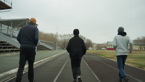 back view of an adult and two young boys jogging side by side on a track in a stadium, with a yellow-painted building in the background, along with parked cars and a grassy field