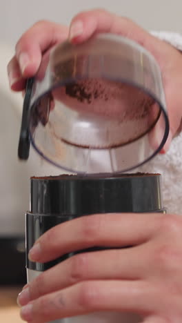 mujer termina de moler granos de café en la mesa de primer plano. dama con bata de baño toca la tapa del molinillo para sacudir el polvo marrón en casa. bebida con cafeína