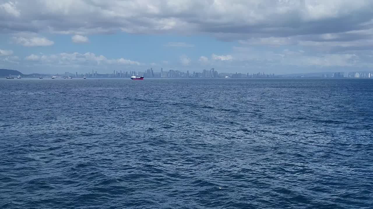 Multiple commercial ships float on the ocean near Panama City, with the skyline visible in the background under a cloudy sky.