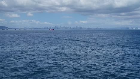 multiple commercial ships float on the ocean near panama city, with the skyline visible in the background under a cloudy sky.