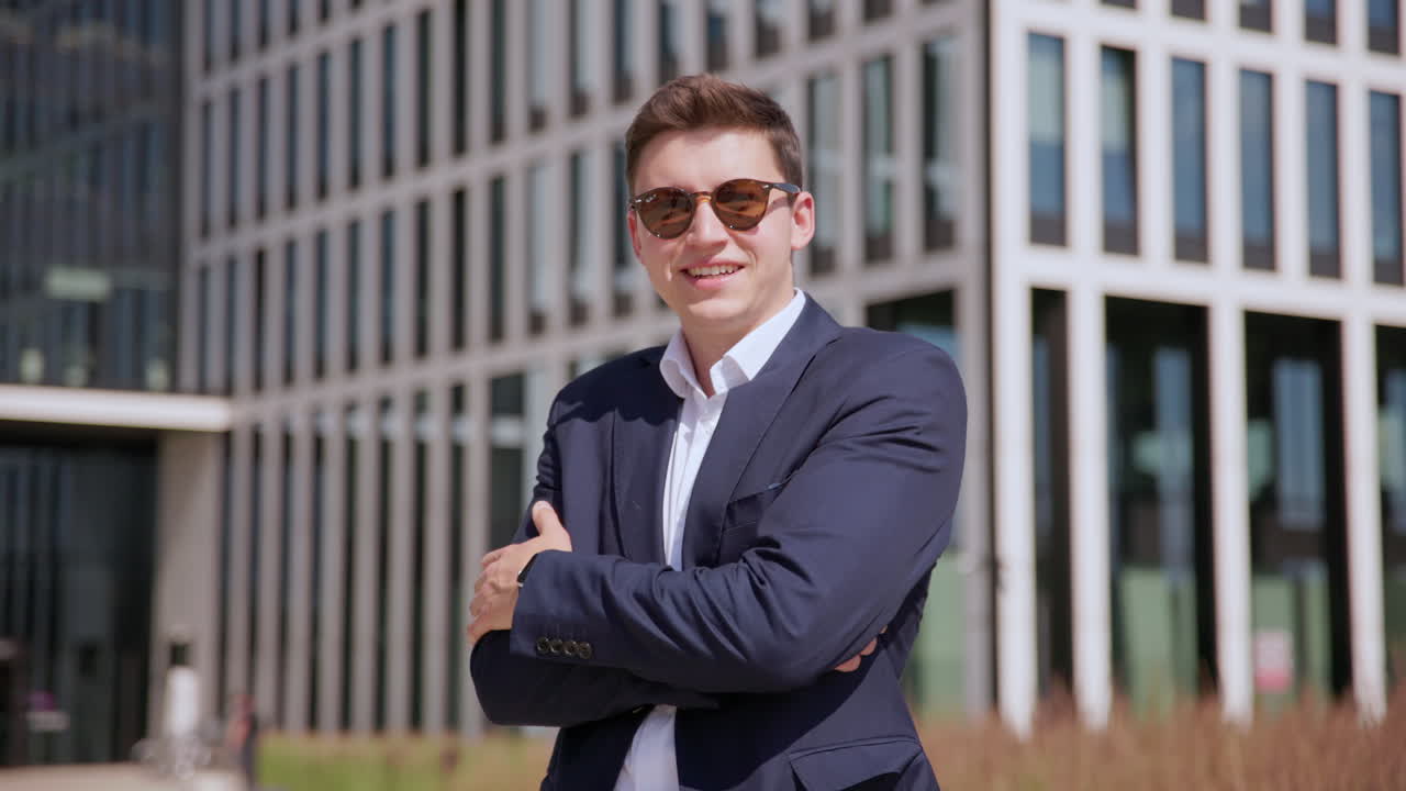 un joven empresario exitoso con gafas de sol se para y sonríe a la cámara frente al moderno edificio de oficinas