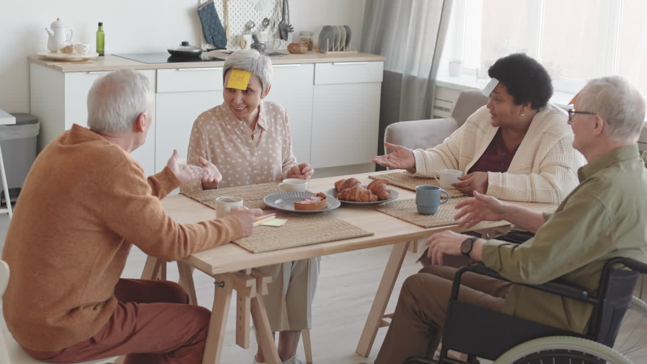 Inquadratura media di diversi amici anziani multietnici con post-it sulla fronte, seduti al tavolo da pranzo, che sorridono, parlano, con una donna asiatica che vince e tutti che applaudono