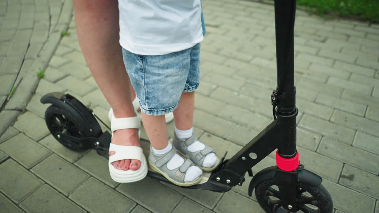 Leg view of a child standing on a black scooter while a woman s foot is also on the scooter, the child has dropped one leg off the scooter