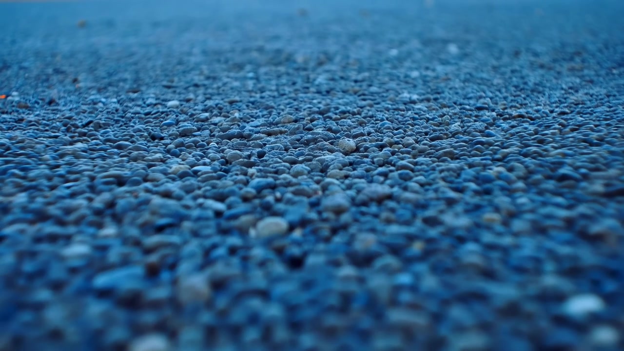 Near-ground panoramic motion across pebble-covered shoreline showing narrow focus plane moving over dense field of small rounded stones, sharper mid-foreground pebbles contrast with softer