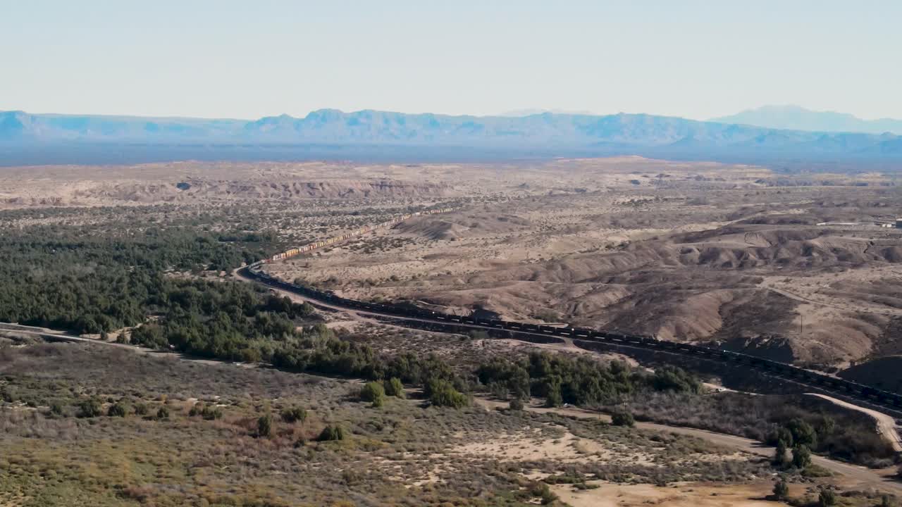 Longo trem de carga transportando mercadorias pelas montanhas do deserto, fronteira entre Califórnia e Arizona, Rio Colorado, autoestrada I-40 Leste, plano aéreo cinematográfico de grande angular de estabelecimento.