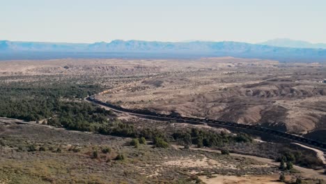 Largo-Tren-De-Carga-Que-Transporta-Carga-A-Través-De-Las-Montañas-Del-Desierto,-Frontera-Entre-California-Y-Arizona,-Río-Colorado,-Autopista-I-40-Este,-Toma-Aérea-Cinematográfica-De-Gran-Angular