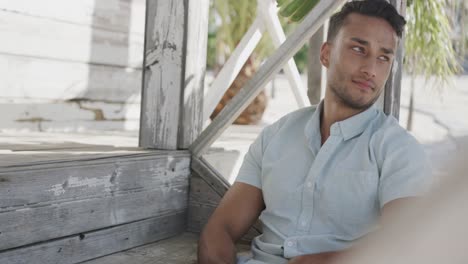 thoughtful biracial man using smartphone sitting on porch of wooden beach house, in slow motion