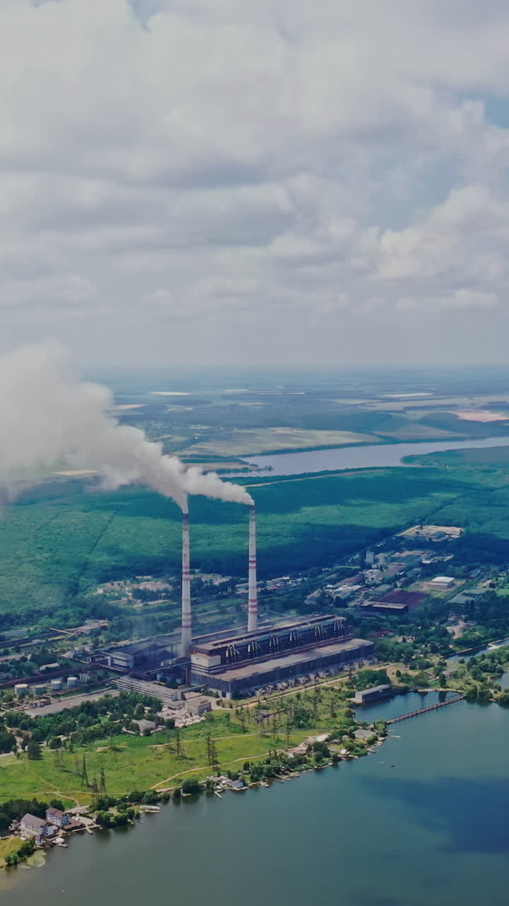 Huge manufactory plant among natural background. Smoke comes from pipes into the atmosphere near the river and green forest outdoors. Aerial view. Vertical video