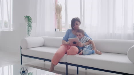 Madre-E-Hijo-Usando-Una-Tableta-Mirando-La-Pantalla-Y-Hablando-En-Un-Sofá-En-Una-Casa-Acogedora