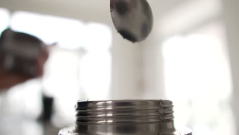 Pouring-ground-coffee-with-metal-spoon-into-silver-coffee-pot