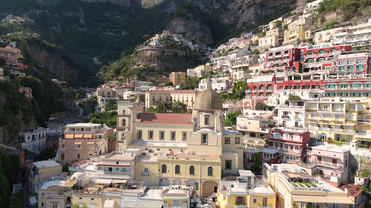 La telecamera cattura una vista dall'alto di Positano, mostrando case ed edifici arroccati sulle colline. Le persone camminano per le strade sottostanti mentre il sole splende luminoso.