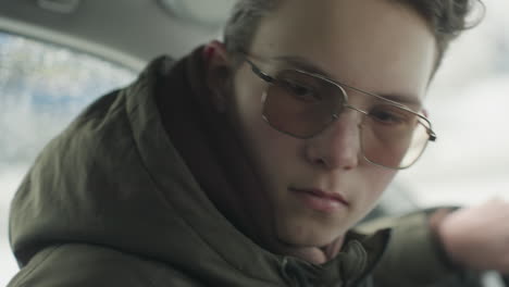 young male driver in sunglasses and green winter jacket turns back inside parked vehicle, beginning to smile while looking directly at camera with soft focus winter background seen through window