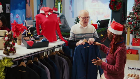 El-Gerente-De-Una-Tienda-Afroamericana-Vende-Un-Blazer-Elegante-A-Un-Cliente-Anciano-Rico,-Convenciéndolo-De-Que-La-Prenda-Está-Magistralmente-Elaborada-Con-Materiales-De-Calidad-En-Una-Tienda-De-Ropa-Con-Adornos-Festivos.