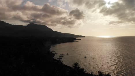 Antena-De-Drones-De-4k-Sobre-Una-Exótica-Isla-Tropical-Paradisíaca-En-El-Océano-Pacífico-Al-Atardecer-Y-Los-Rayos-Del-Sol-Brillando-Intensamente-A-Través-De-Un-Cielo-Nublado-Y-Un-Pico-De-Volcán-Y-Una-Costa-Oscura-Con-Barcos-En-El-Mar